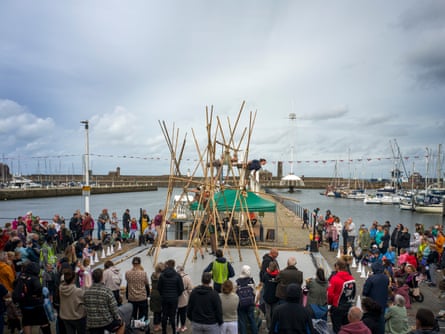 A crowd of people watch acrobats performing on a temporary structure on a waterfront pier