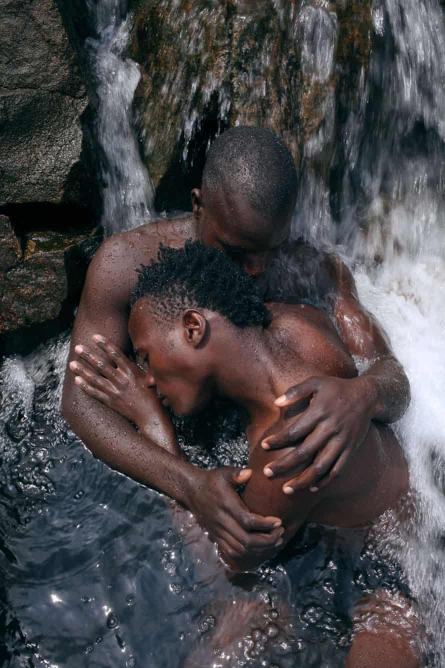 One nude young Ugandan man embraces another as they sit under a waterfall, in a pool of water. Their eyes are closed, and their heads are tilted away from the camera.