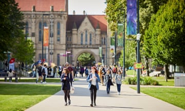 Students walking on a wide path outside in the sun on a university campus, with grass and trees outside and a building in the background
