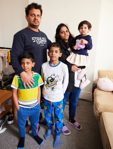 Geo Ambooken, his wife, and their three young children, all standing in a living room with a pale brown sofa, wooden table and grey carpet. Children’s clothes are seen hanging on a drying rack and shoes under the table. The mother is holding the little girl in her arms while the two boys stand in front of their father; the boys are dressed in T-shirts and pyjama trousers.