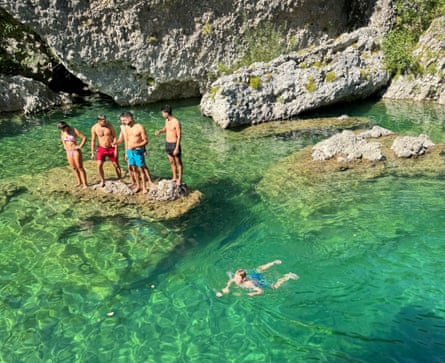 Five people standing on a rock in a natural pool surrounded by rocks, and one person swimming.