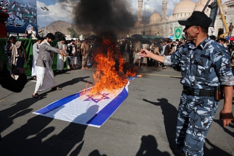 A uniformed officer watches as a crowd of people gather around a burning Israeli flag on the ground.