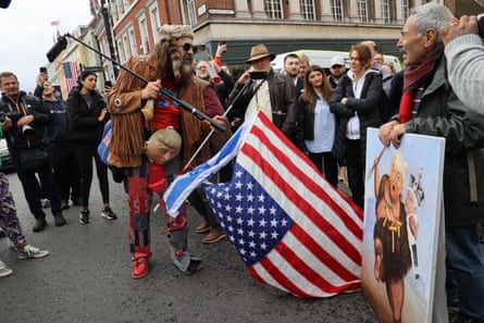 A bearded protester in a brown fringed suede jacket, coonskin cap and Trump mask around his waist holds an Israel flag in one hand and a selfie stick in another