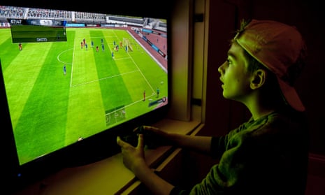 A boy with a baseball hat on backwards plays a football game with his games console.