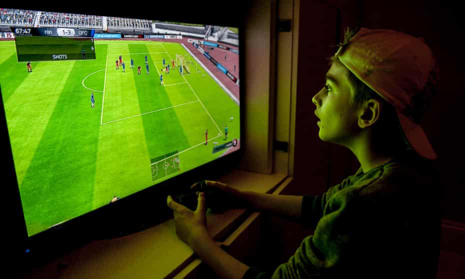 A boy with a baseball hat on backwards plays a football game with his games console.