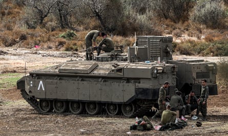 a group of Israeli soldiers sit, stand and lie on the ground by a tank, several talking on phones; they look relaxed; two more soldiers are on the tank and appear to be adjusting or checking the gun turret