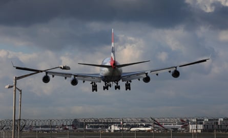 Plane landing at Heathrow airport, seen in closeup from behind; the British Airways colours are seen on its tail. It is framed against a dark grey, cloudy sky.