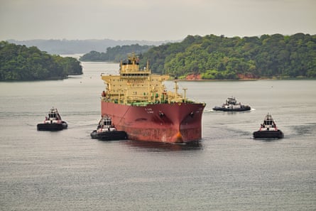 A large oil tanker surrounded by four tug boats on the Panama Canal, with rainforest on the shore.