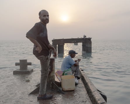 Man stood, another seated on small jetty at sea with sun in background