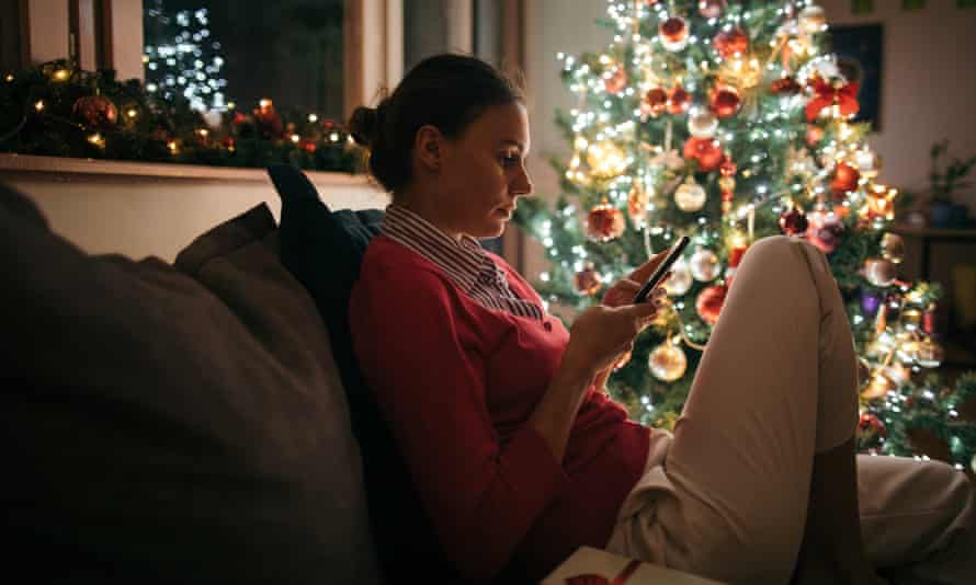 A woman sitting on a couch in front of a lit-up Christmas tree, looking at her phone