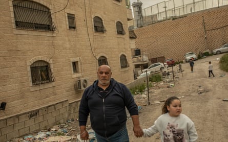 Ali Halhouli stands holding the hand of a young girl on a dusty road outside a residential building. At the end of the road behind is a large wall with wire fencing on top, and behind that is the watchtower