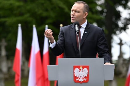 Karol Nawrocki speaks and gestures at a lectern outdoors with flags and trees behind him