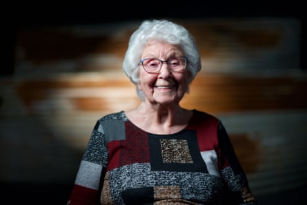 A woman with white hair, smiling, wearing a grey, black and red jumper with geometric shapes.
