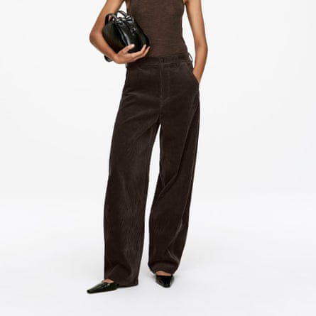 A model wears dark brown Barrel-Leg Corduroy Trousers from Arket. They wear flat point shoes, a sleeveless brown top and are holding a handbag.
