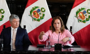 Woman dressed in pink suit holds necklace and sits with man in front of Peruvian flags