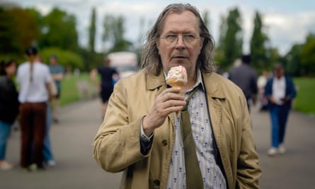 Gary Oldman, dishevled in an overcoat and tie, with greasy long hair, walks in a park eating an ice-cream cone