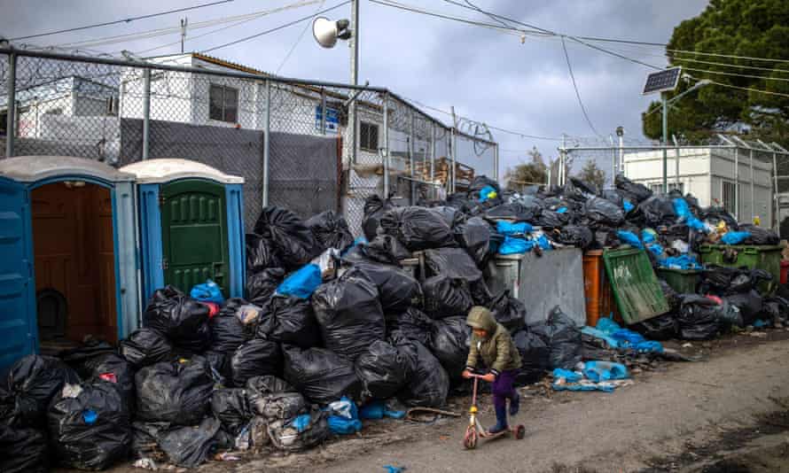 A boy plays with a scooter in front of rubbish bags in area outside camp Moria.