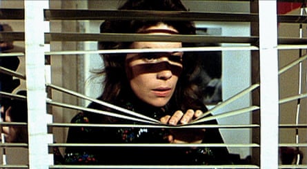 Jennifer Salt holding down and looking through a set of venetian blinds which frame her shadowy face