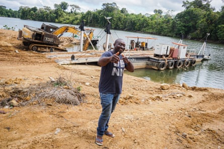 A man smiles and points at the camera. Behind him is a river, a small ferry and an excavator.
