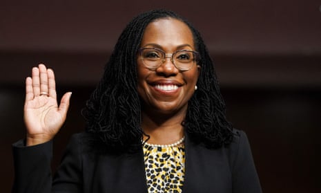 Ketanji Brown Jackson is shown smiling as she raises her right hand to take an oath. She has a microlocked hair style that flows just past her shoulders and is wearing glases, a black suit and a black, white and yellow patterned blouse.