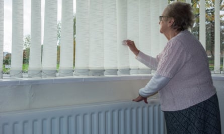 An elderly woman stands with a hand on a radiator in her house