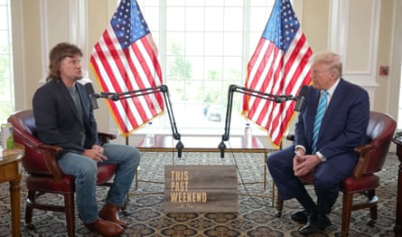 Two men sit in front of microphones with two US flags as backdrops for a podcast interview