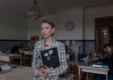 A young woman holding a folder in front of her stares into the distance in an office with three other women at desks working on computers behind her