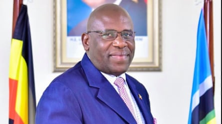 Portrait of Asuman Kiyingi wearing suit and glasses, standing between two flags