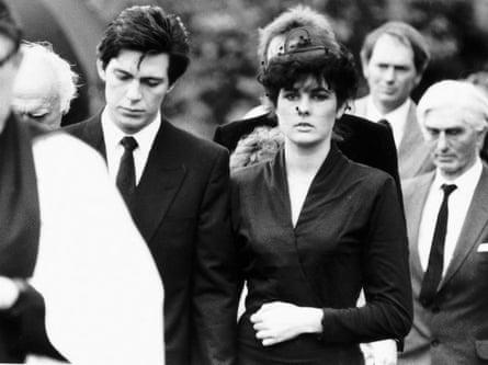 A funeral gathering. A man in a dark suit and tie with a woman in a black suit and black hat