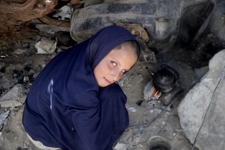 A young girl in blue robe and headscarf crouches down with a teapot on a fire