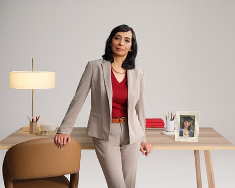 Smitha, an MBA graduate, leans against a wooden desk in a beige suit, smiling