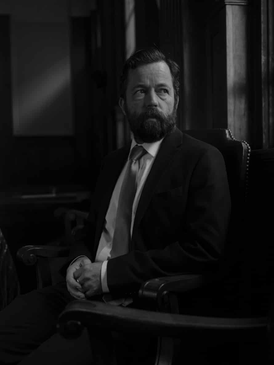 A man with a beard and mustache wearing a suit and tie sits for a portrait in a court room