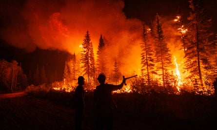 Two men in helmets silhouetted against huge flames on a wooded hillside