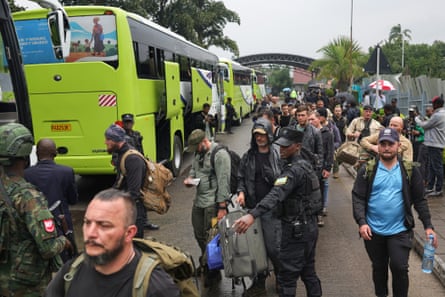A large group of men walk past three green coaches, watched by military guards