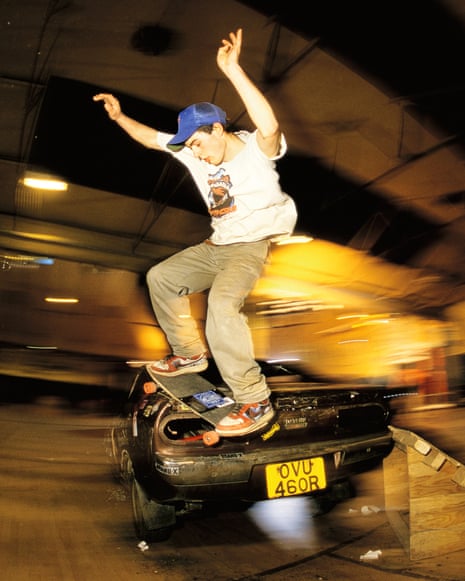 A skateboarder mid-trick above a paved area, with a red car and weathered buildings in the background