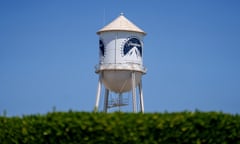 A white water tower with the dark blue Paramount logo on it, of a mountaintop surrounded by stars, rises beyond a green hedge blurry in the foreground.