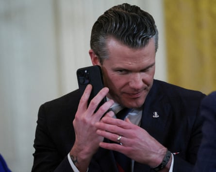 A besuited man, Pete Hegseth, holds a phone with both hands like it’s a precious object