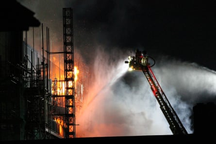 Firefighters on a cherrypicker ladder spray water on a burning building with scaffolding around it