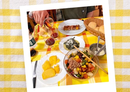 A table spread with salad, grilled meat skewers, polenta, greens, bread, and wine on a checkered tablecloth