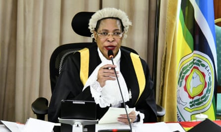 An African woman in an 18th-century white wool wig, lace collar and gown sits at a desk beside a flag  