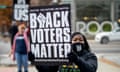 A young Black woman wearing a black face mask and sweatshirt holds a white-on-black sign that says Black Voters Matter.