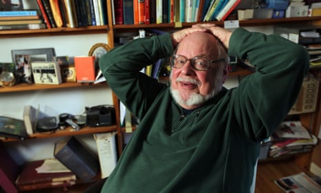 Norton Juster leaning back in his chair with his hands clasped on his head, in a green jumper, with shelves full of books behind
