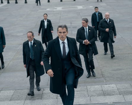 A dramatic wide shot from The Diplomat shows actor Rufus Sewell as Hal Wyler walking purposefully forward in a wide, gray paved courtyard.