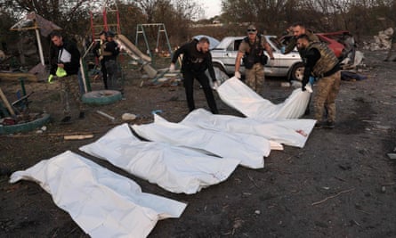 Officers lay down a white body bag on the ground next to four others with cars and playground equipment in the background.