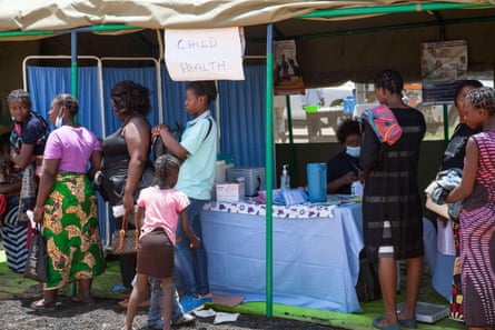 people stand in a line in front of a temporary structure bearing a hand written sign that reads 'child health'.