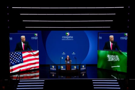 wide view of a man in a suit speaking into a microphone on stage flanked by two screens showing his image and the US and Saudi flags