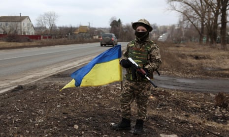 A boy wearing military-camo clothes holds a toy machine gun next to a fluttering Ukrainian flag on a road in Korobochkino village.