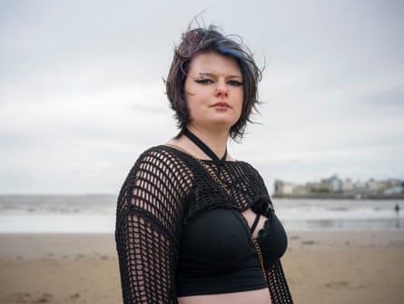 A young woman in a black crop top, face studs, and streaked blue hair stands on a beach
