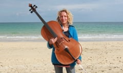 Writer Catrina Davies, holding her cello, while stood on a sandy beach on a sunny day.
