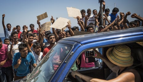 A woman takes a picture from her car of a group of Pakistani refugees protesting at their lack of progress obtaining transit papers in Kos.